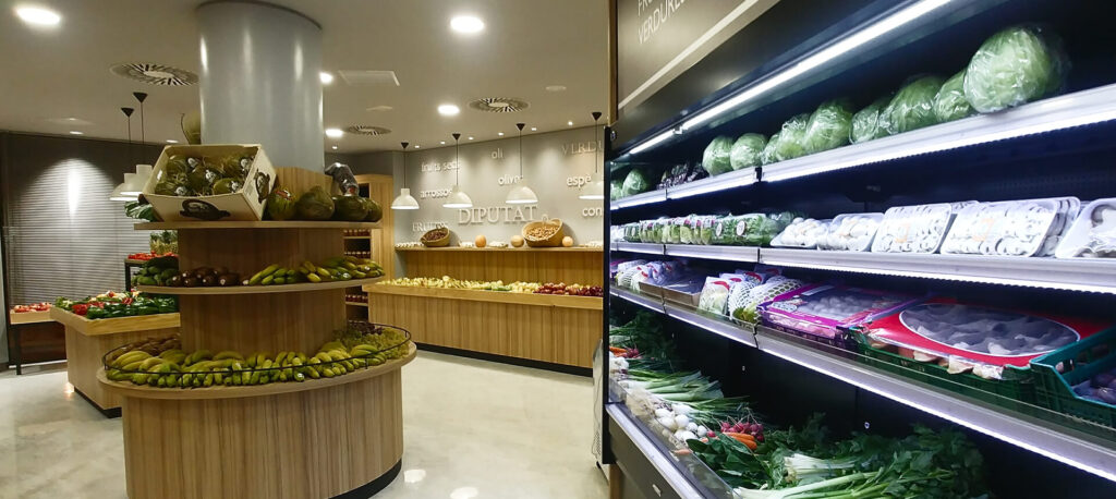 Interior de supermercado con zona de frescos, expositores de fruta y verdura y mural refrigerado con producto envasado.
